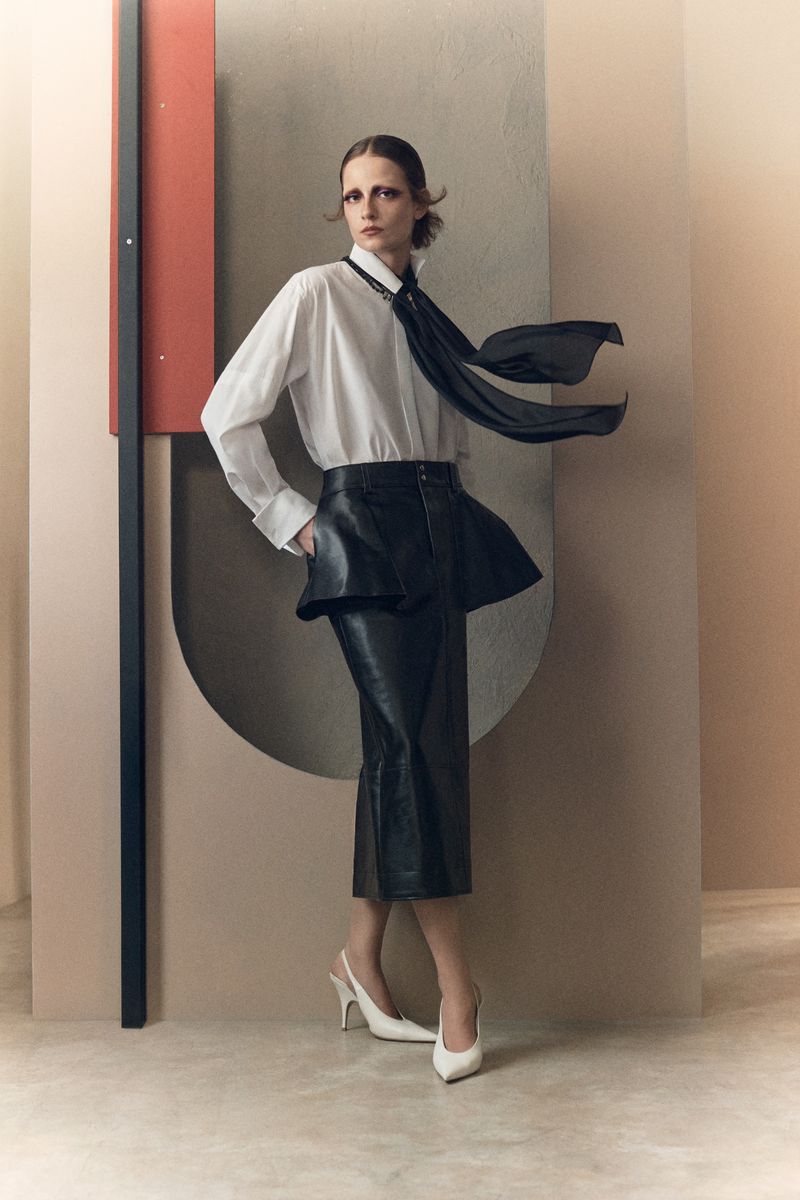 Model in white shirt with black tie, black leather peplum skirt and white heels against structured set backdrop