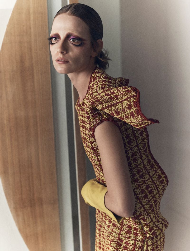 Model in patterned red and yellow dress wearing yellow gloves against a minimalist interior background.