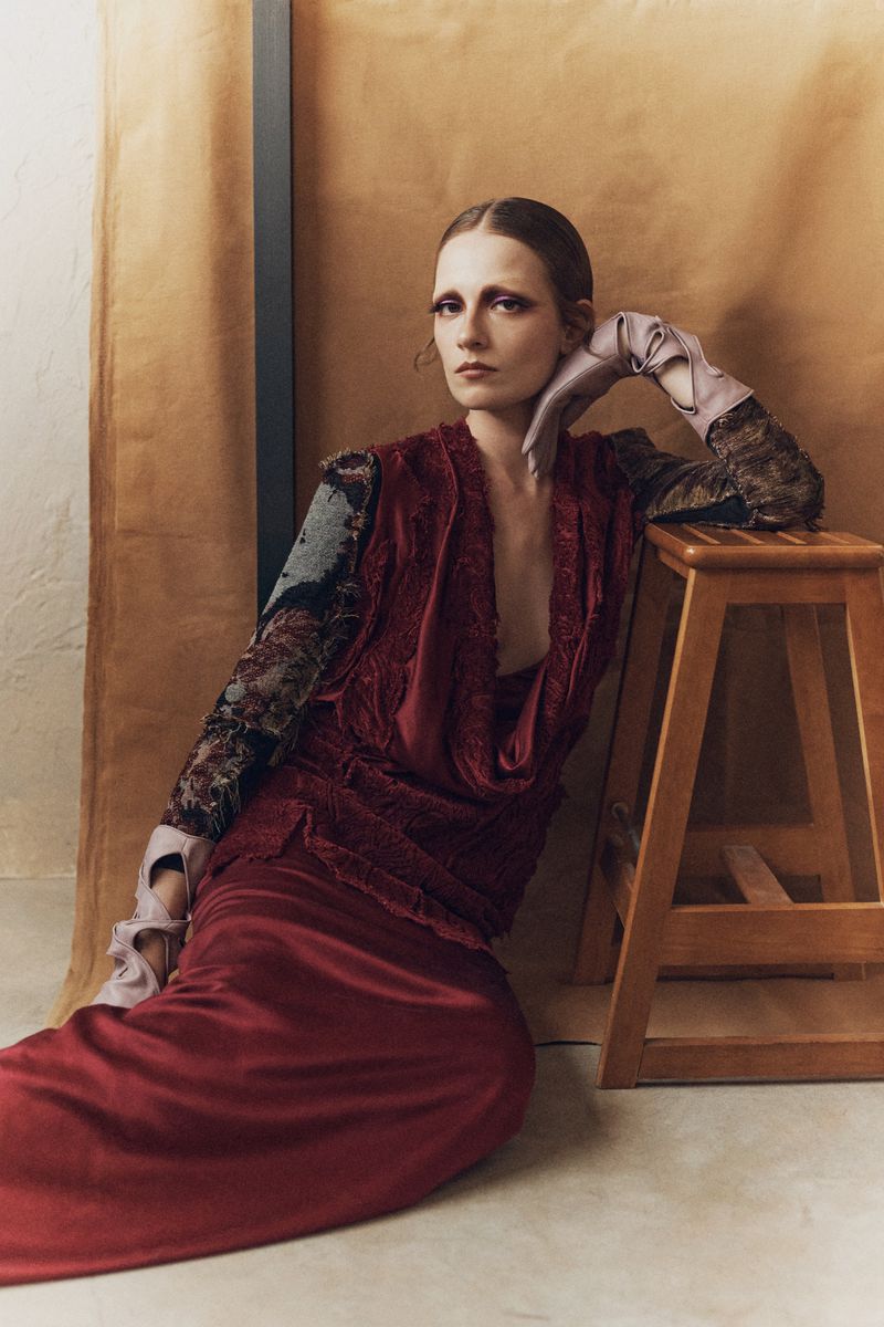 Model in burgundy silk dress and pink leather gloves leaning on a wooden stool against a textured backdrop