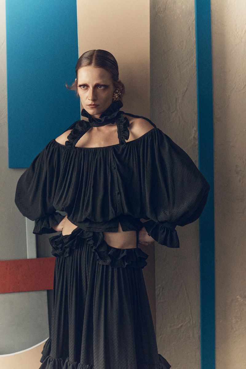 Model in ruffled black top and skirt posing against geometric multi-toned panels for Vogue Greece