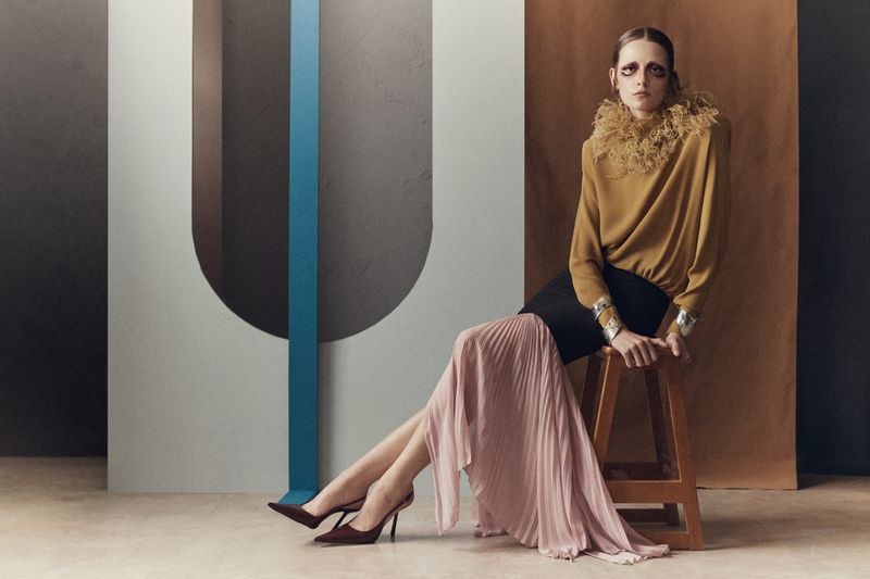 Model seated on wooden stool wearing ruffled yellow top and pink pleated skirt against studio backdrop