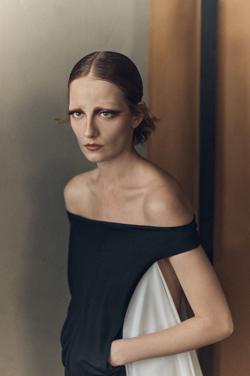 Model with slicked-back hair and bold eye makeup wearing a black off-the-shoulder dress for Vogue Greece.