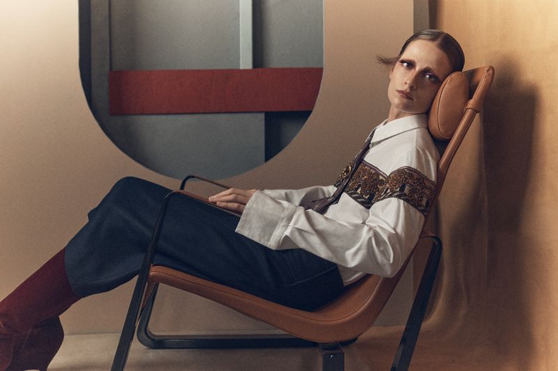 Model Sophie Dominique Gonzalez seated in a leather lounge chair wearing an embroidered white shirt and dark trousers against a geometric set backdrop