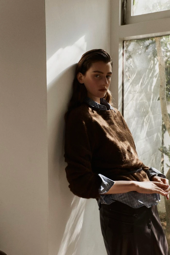 Model leaning against a white wall next to a window wearing a brown sweater over a patterned shirt for Lula Japan