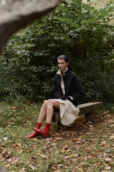 Model seated on a wooden bench outdoors wearing a dark jacket, cream skirt, and vibrant red socks with shoes.