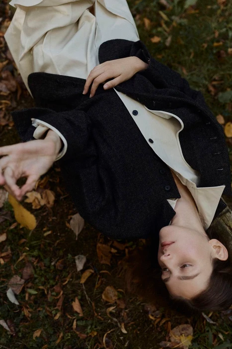 Model lying on forest ground wearing a dark jacket and white shirt surrounded by fallen leaves