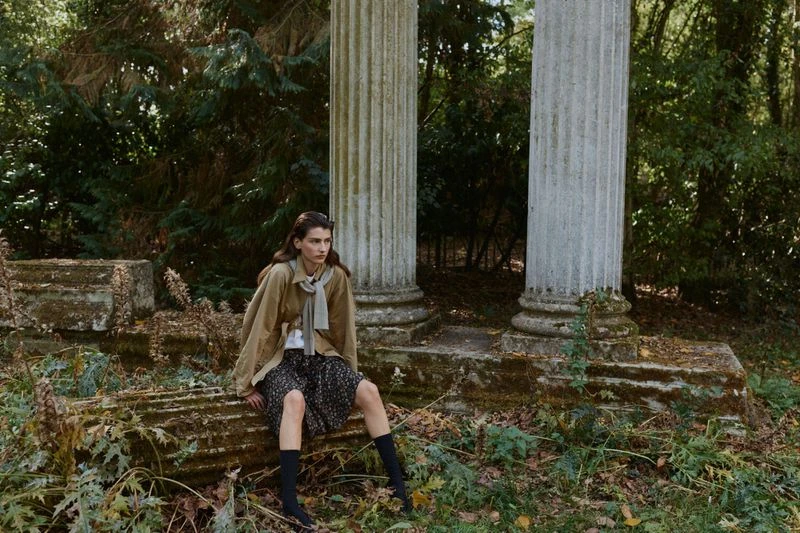 Model seated on stone ruin among classical columns and overgrown foliage wearing a beige jacket and patterned skirt
