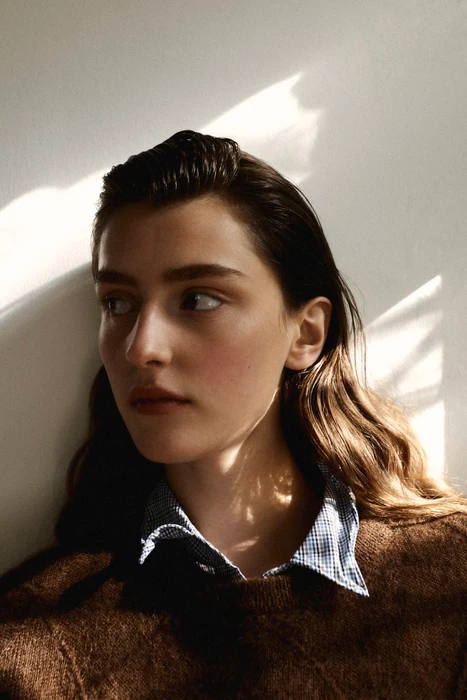 Portrait of a woman wearing a brown knitted sweater and a blue-and-white checked collared shirt against a plain wall.