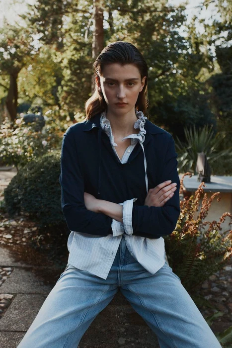 Model standing outdoors with arms crossed wearing a navy blue sweater over a white shirt and blue jeans in a garden setting