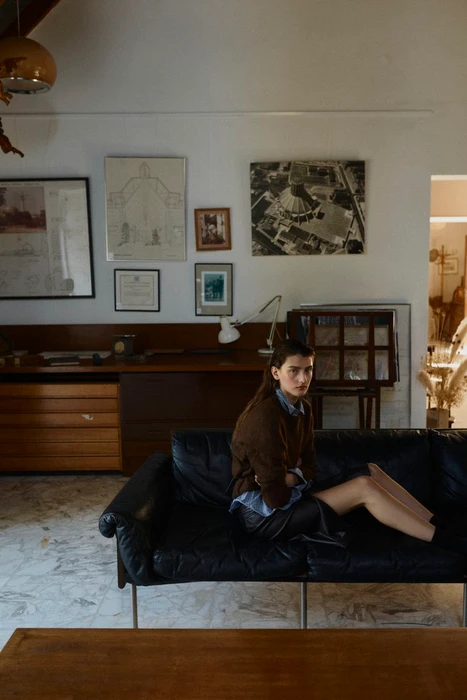 Model seated on a black leather sofa in a domestic living room interior, wearing a brown sweater and collared shirt