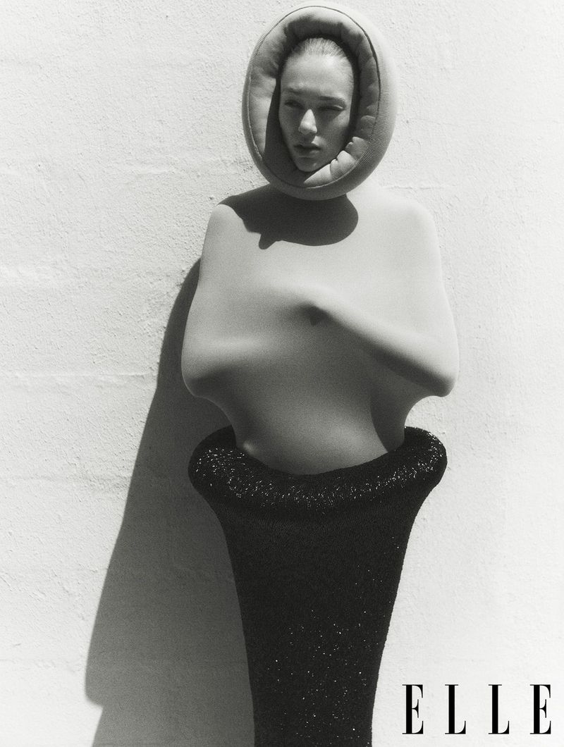 Black and white fashion shot of a woman in a sculptural garment against a textured wall. 'Elle' logo visible in the lower right.