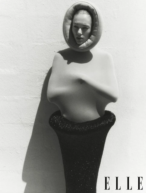Black and white fashion shot of a woman in a sculptural garment against a textured wall. 'Elle' logo visible in the lower right.