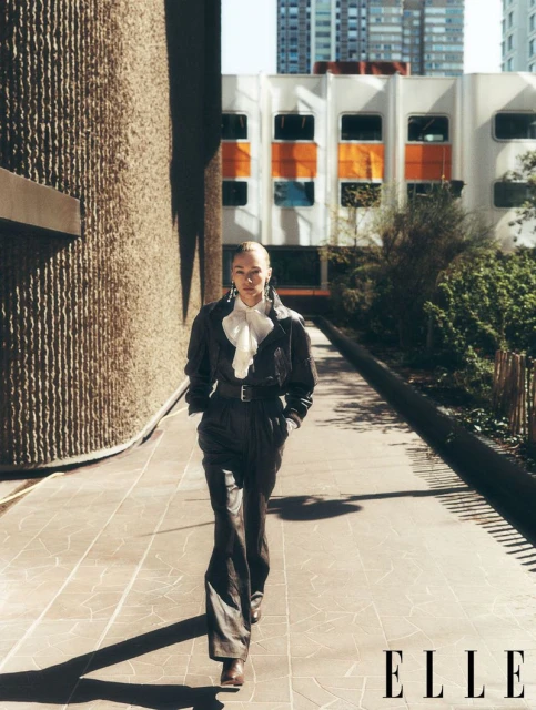 Stylish adult woman in a dark leather jumpsuit walks on a tile path near brutalist architecture in urban daylight. Fashion editorial.