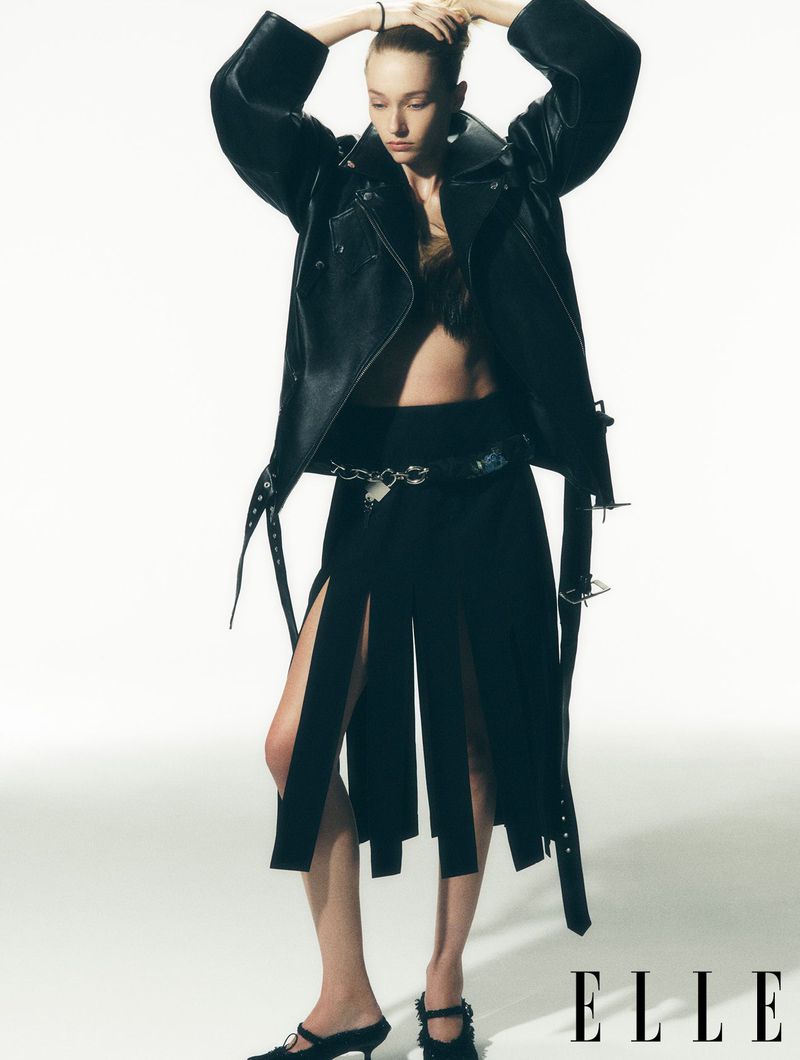 Editorial fashion shot of a slender woman in a black leather jacket and deconstructed skirt, posing against a white background in a studio.