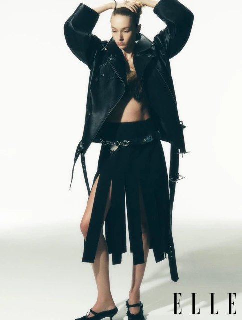 Editorial fashion shot of a slender woman in a black leather jacket and deconstructed skirt, posing against a white background in a studio.