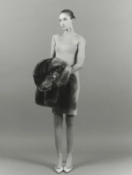Model wearing a short-sleeved fitted dress holding a fluffy stole against a grey studio backdrop