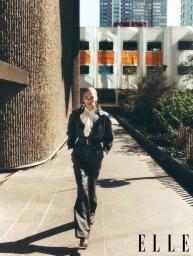Model walking outdoors along a tiled walkway between a textured concrete wall and a modern building.