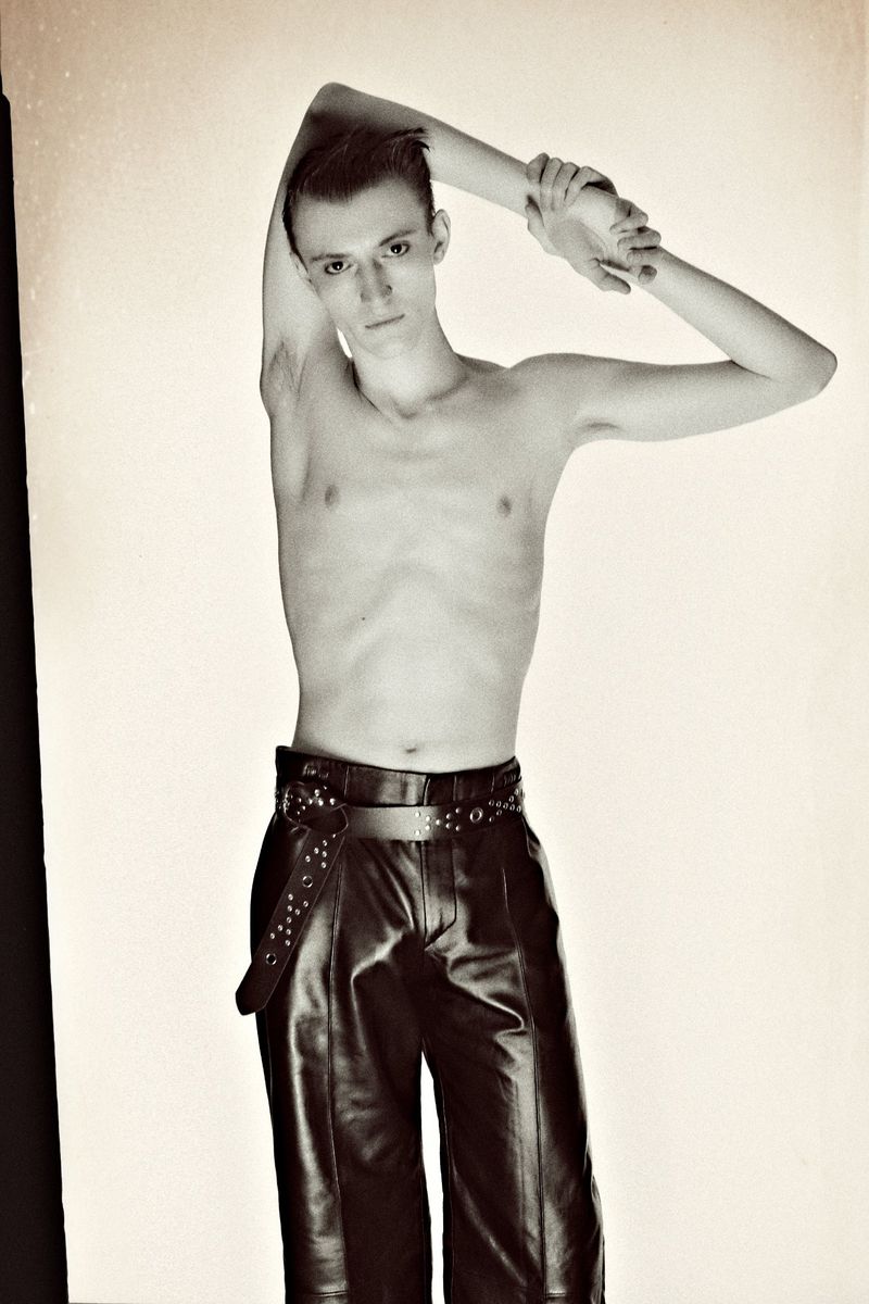 Monochrome studio shot of a slim adult male with arms raised, wearing black leather pants and a studded belt against a white background.