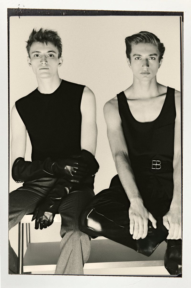 Two men in black tops and dark pants sit side-by-side on a white bench. One wears gloves. Sepia tones, indoor studio setting.