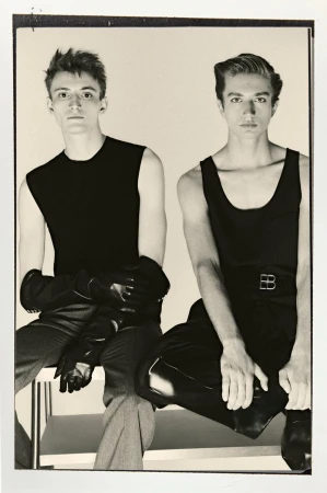 Two men in black tops and dark pants sit side-by-side on a white bench. One wears gloves. Sepia tones, indoor studio setting.