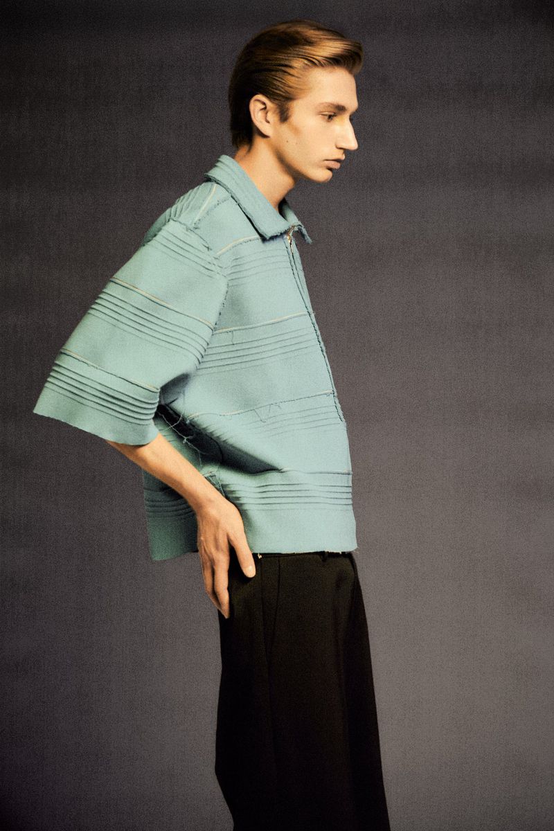 Man in a light blue, horizontal striped shirt and black pants poses in a studio with a neutral gray background in a fashion editorial.