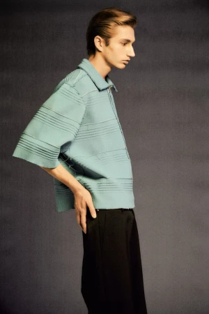 Man in a light blue, horizontal striped shirt and black pants poses in a studio with a neutral gray background in a fashion editorial.