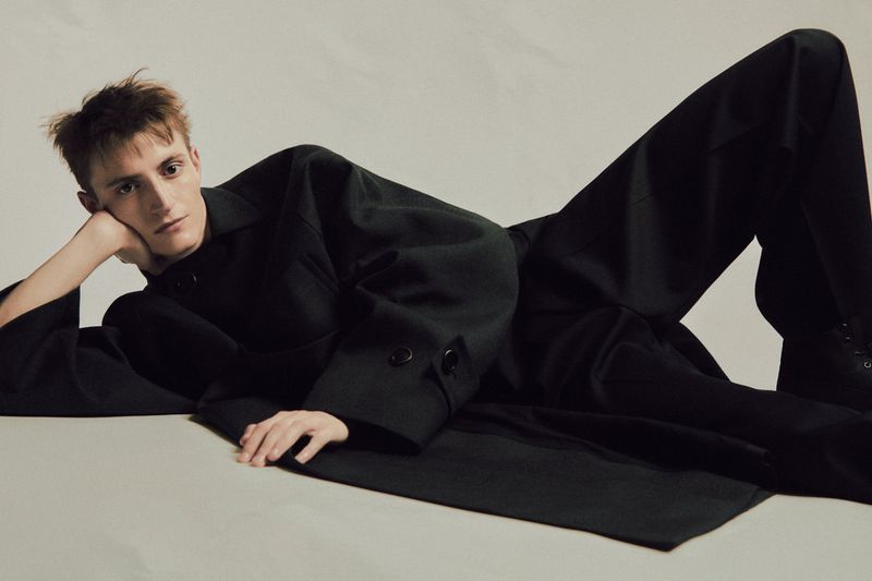 Adult male laying down wearing a black coat. He's resting his head on his hand in an indoor photo studio with soft lighting.
