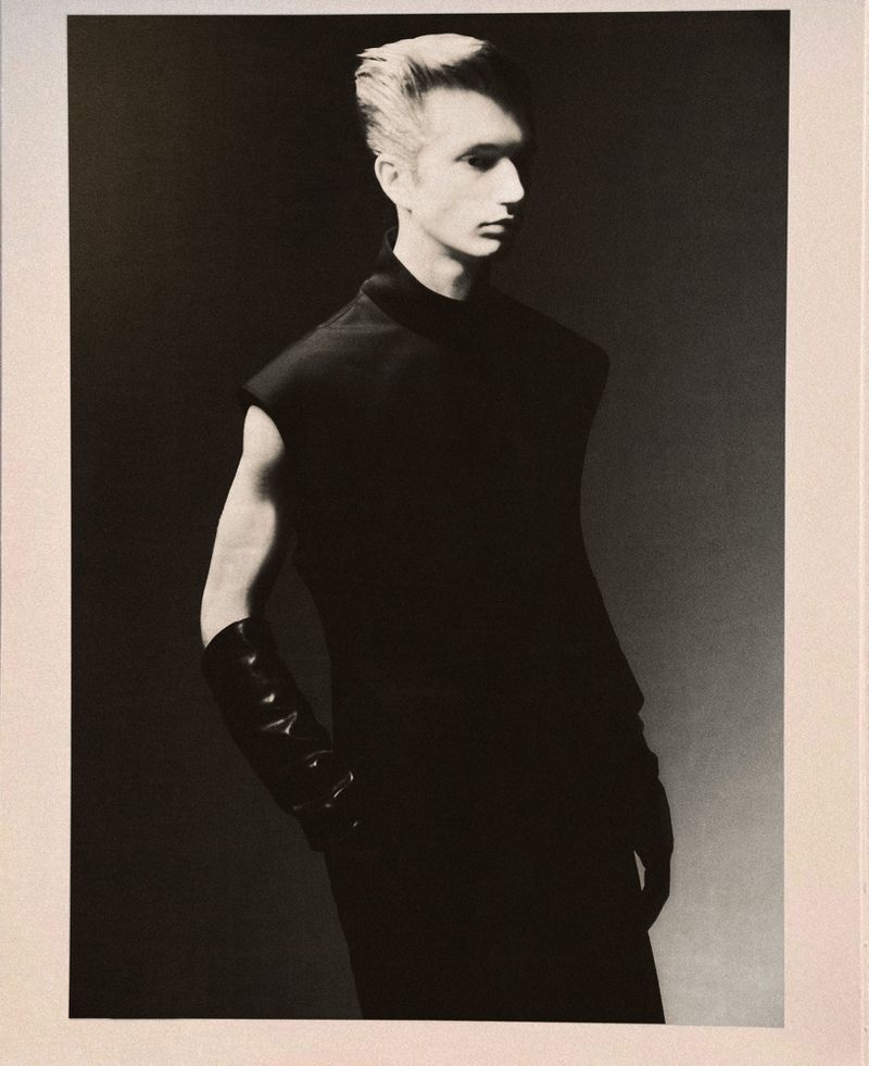 Fashion portrait of a male model in a black sleeveless top and gloves against a grey gradient backdrop, shot in a photo studio.