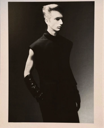 Fashion portrait of a male model in a black sleeveless top and gloves against a grey gradient backdrop, shot in a photo studio.