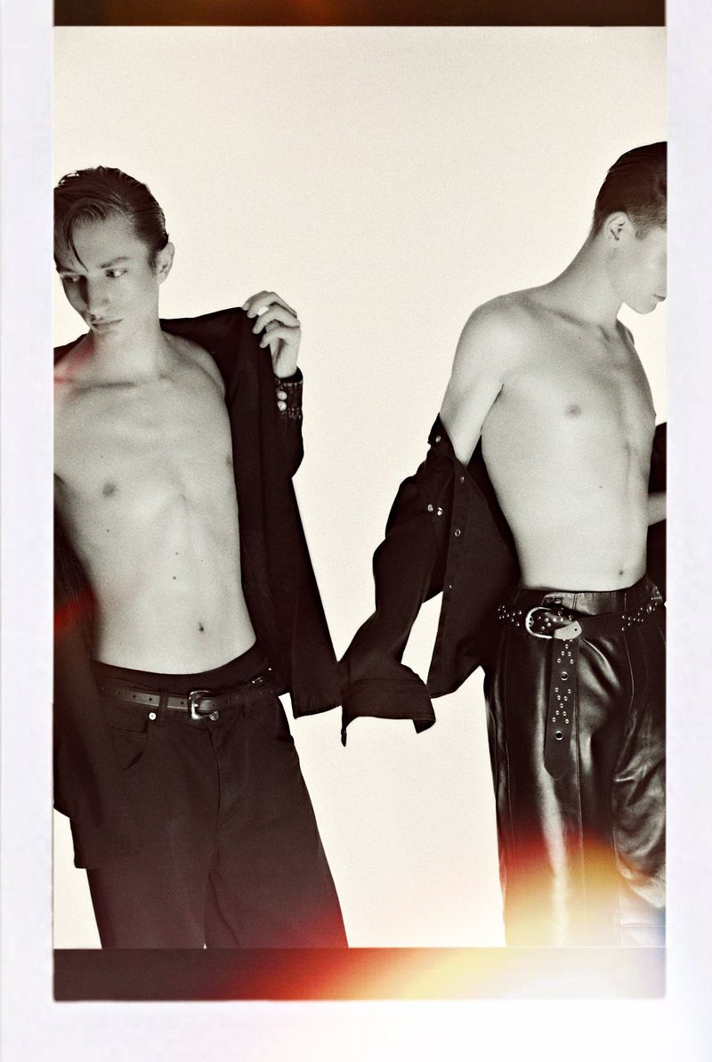 Black-and-white studio shot featuring two adult men, bare-chested, wearing black jackets and pants, standing against a light backdrop.