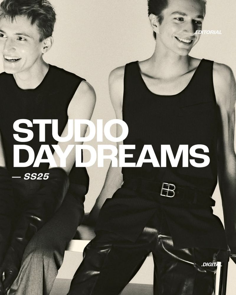 Editorial fashion shot of two slender adult male fashion models in dark tank tops and pants, posing in front of a neutral background.