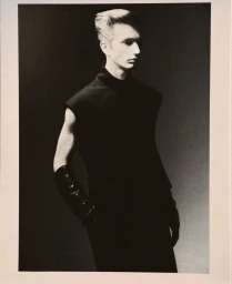 Monochrome portrait of a male model wearing a dark sleeveless top and gloves against a gradient studio background.