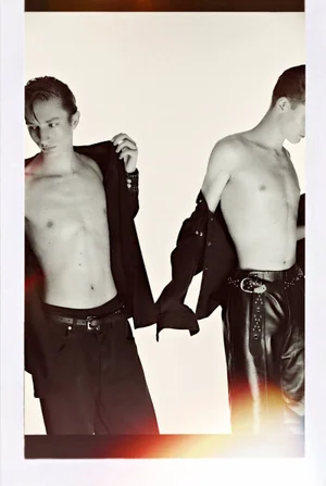 Two shirtless male models wearing dark trousers against a plain white studio background in an editorial fashion portrait.