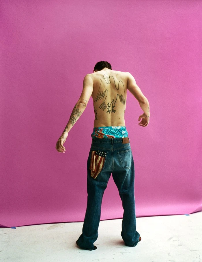 Shirtless man seen from behind with large back tattoo wearing blue jeans and patterned boxers against pink backdrop