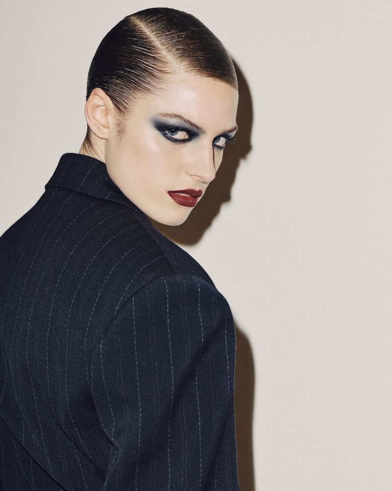 Model in dark pinstripe jacket with slicked hair, dramatic blue eyeshadow and dark red lips against a neutral background