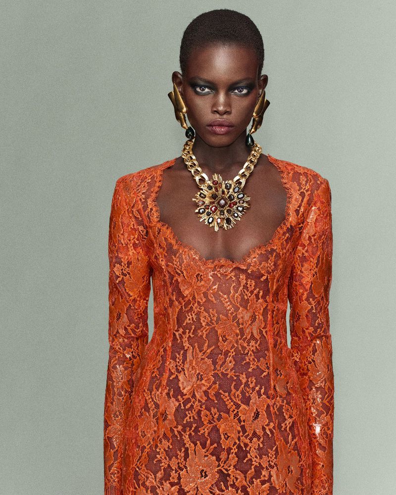 Model wearing a textured orange lace dress with a large gold statement necklace and earrings against a plain muted background.
