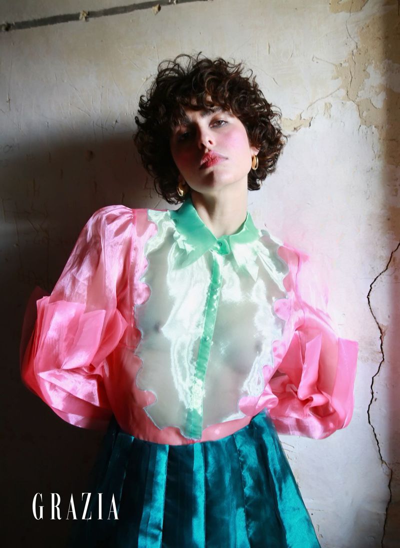 A woman with curly brown hair poses indoors in a fashion editorial wearing a pink and green blouse and a teal skirt.