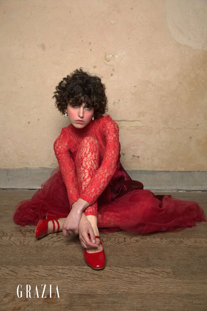 A woman in a red lace dress and patent leather shoes sits in front of a textured wall with the Grazia logo in the corner.