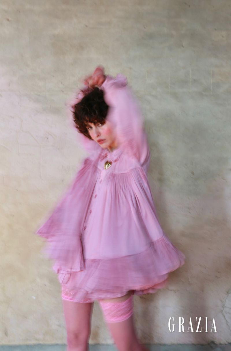 Young woman with dark curly hair poses in a pink vintage-style dress and matching tights in a blurry, soft-focus studio fashion shot.