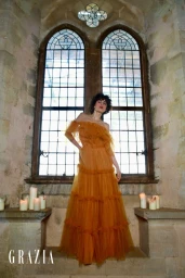 Model standing before arched stained-glass windows in an off-the-shoulder tiered amber dress for Grazia Slovenia