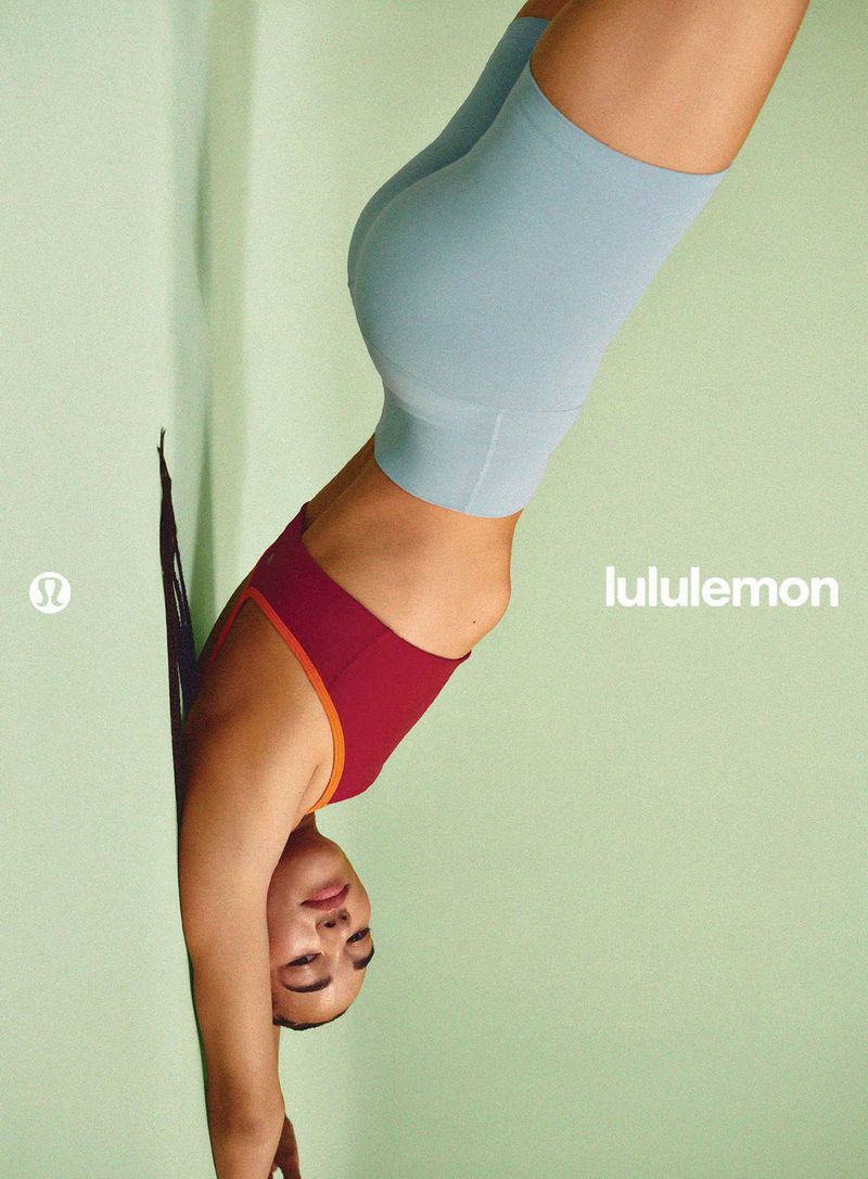 Model in a maroon sports bra and powder blue athletic shorts performing a handstand against a pale green wall.