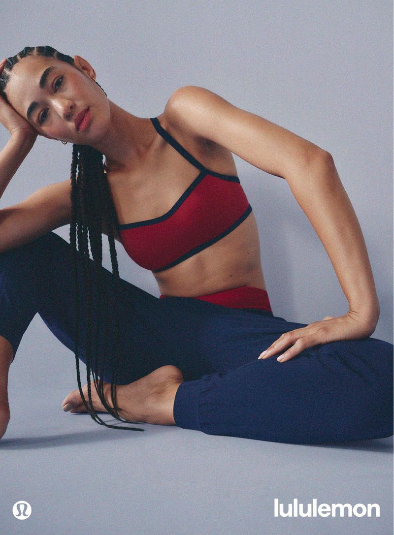 Model with long braided hair wearing a red sports bra and blue pants sitting against a grey seamless background for lululemon