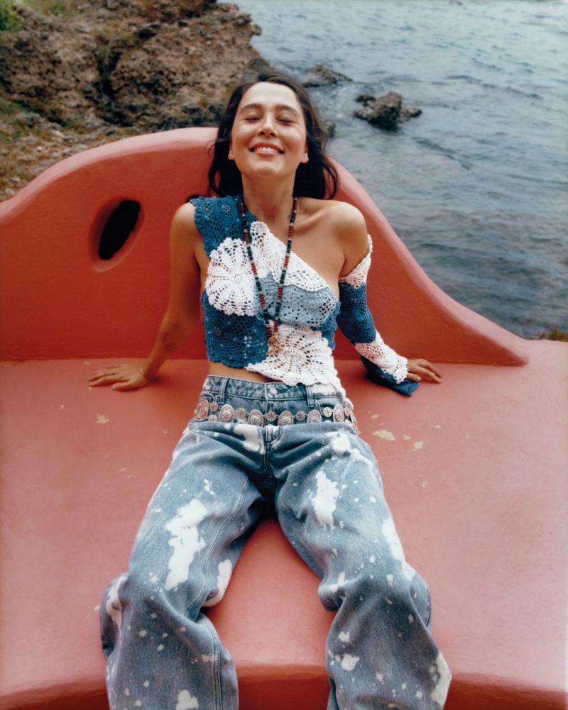 Woman in crochet top and jeans sits on a bench against a coastal backdrop, radiating joy with her eyes closed and a wide smile.