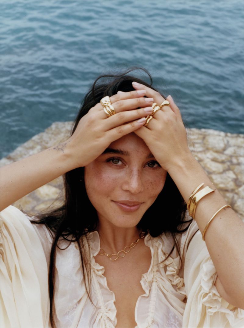 Portrait of a smiling woman wearing gold jewelry and a cream ruffled blouse against a coastal ocean backdrop.