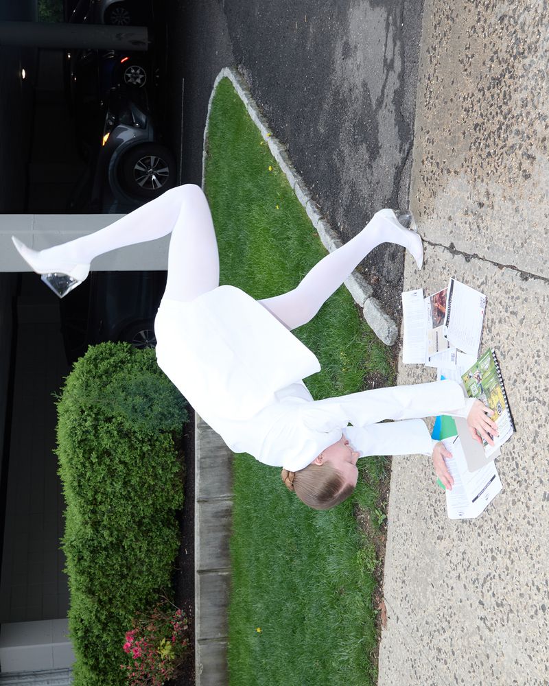 Woman in white suit and tights lying on pavement surrounded by papers and books, with green bushes and a car in the background.