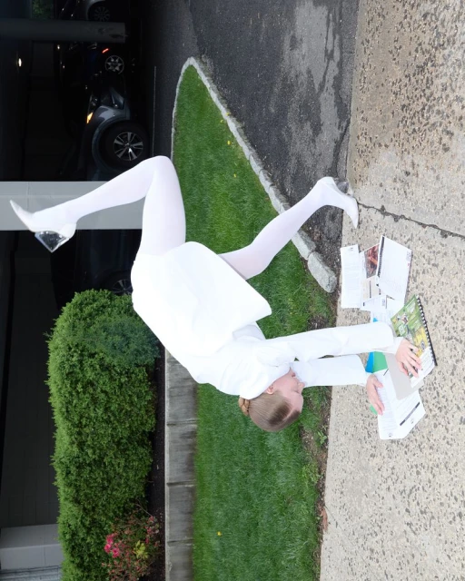 Woman in white suit and tights lying on pavement surrounded by papers and books, with green bushes and a car in the background.