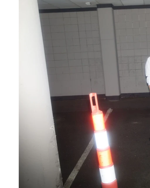 Indoor setting features an orange and white safety cone on a dark floor, a gray wall, and a white column in a partially lit space.