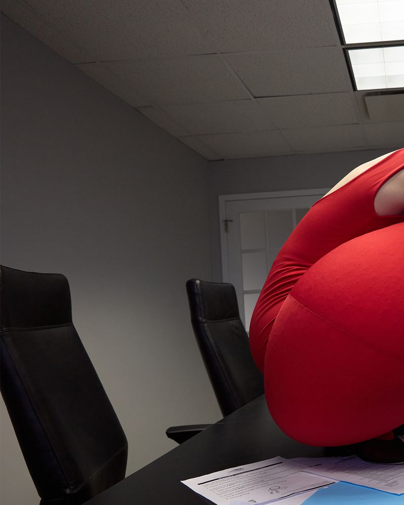 Adult in red leotard sitting on a black desk with papers and a blue folder in an office setting with black office chairs.