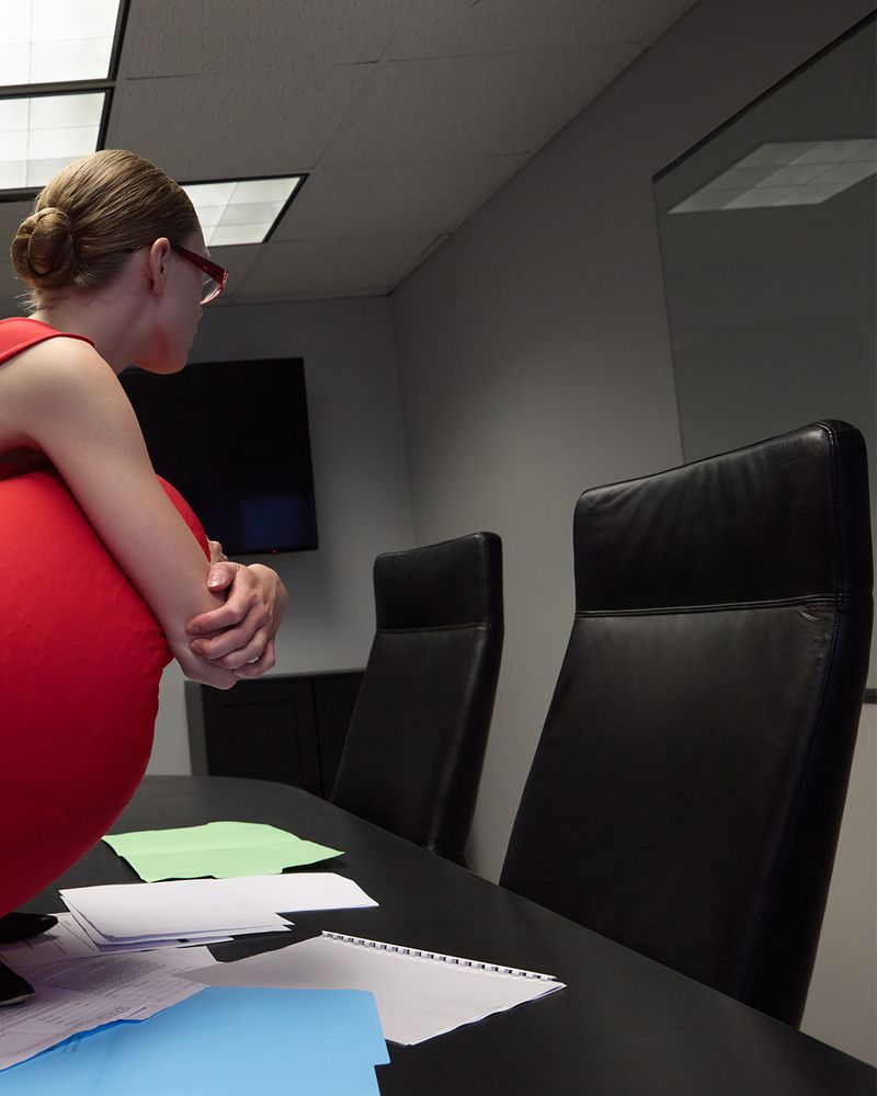 Adult woman in red dress crouching on a table in an office environment with papers and chairs, creating a modern business scene.