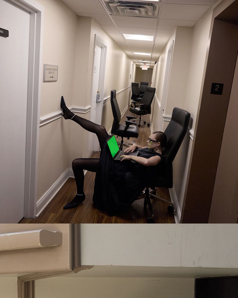 Woman in black dress sitting in an office chair working on a laptop in a hallway with office chairs; workplace.